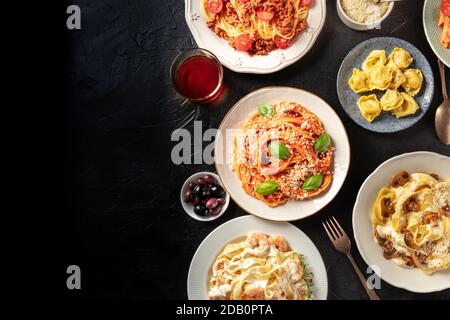 Italienisches Essen, überhängender Flachschuss. Pasta, Oliven, Wein auf schwarzem Hintergrund mit Kopierraum. Spaghetti mit Tomatensauce, Ravioli und anderen Stockfoto