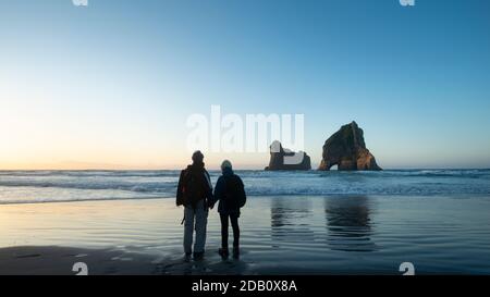 Paar stehen am Wharariki Beach und beobachten Archway Island bei Sonnenuntergang, Südinsel von Neuseeland Stockfoto