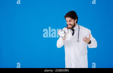 Ein junger bärtiger Arzt auf blauem Hintergrund mit einem Megaphon in seinen Händen schreit Stockfoto
