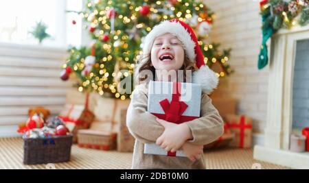 Frohe Weihnachten und schöne Feiertage! Fröhliche niedliche Mädchen mit Geschenk. Kind hat Spaß in der Nähe von Baum. Stockfoto