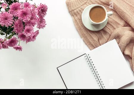 Gemütlicher weiblicher Arbeitsplatz mit leerem Notizbuch, Stift, Tasse Kaffee, Strickpullover und einem Blumenstrauß Chrysantheme. Weißer Hintergrund, Draufsicht, f Stockfoto