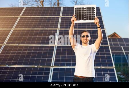 Stilvoller Kerl hält ein Solarpanel über sich. Im Hintergrund befinden sich Reihen von Solarzellen in den angrenzenden Bereichen. Ein Mann in Sonnenbrille und einem weißen T-Shirt lächelt, während er auf die Kamera schaut. Stockfoto