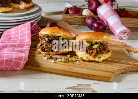 Hausgemachter Pulled Beef Burger mit karamellisierten roten Zwiebeln, Gurken und Käse auf einem Holzbrett Stockfoto