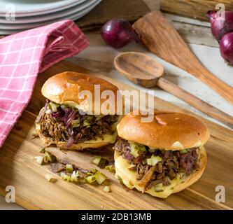 Hausgemachter Pulled Beef Burger mit karamellisierten roten Zwiebeln, Gurken und Käse auf einem Holzbrett Stockfoto