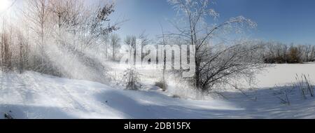 rime Eis und Schnee auf den Ästen von Bäumen am Ufer des gefrorenen Sees, die Strahlen der Morgensonne durch die Bäume Stockfoto