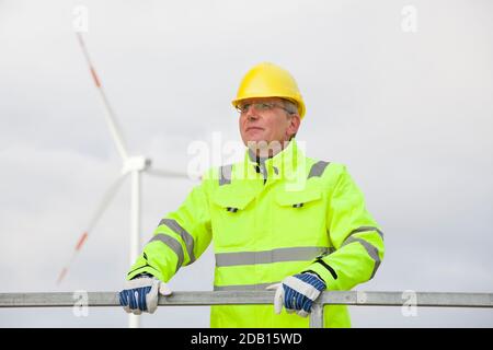 Reifer Ingenieur mit Hut und Schutzkleidung vorne Der verschwommenen Windenergieanlage im Hintergrund Stockfoto