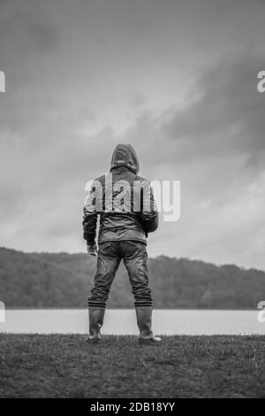 Porträt, mono, Rückansicht eines isolierten Mannes, der in Gummistiefel steht, Mantel mit Kapuze und Blick über Wasser mit launischen Wolken darüber; bedrohliches Aussehen. Stockfoto