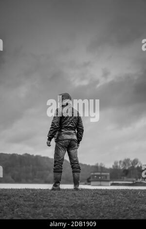 Porträt, mono, Rückansicht eines isolierten Mannes, der in Gummistiefel steht, Mantel mit Kapuze und Blick über Wasser mit launischen Wolken darüber; bedrohliches Aussehen. Stockfoto