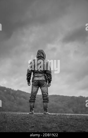 Porträt, mono, Rückansicht eines isolierten Mannes, der in Gummistiefel steht, Mantel mit Kapuze und Blick über Wasser mit launischen Wolken darüber; bedrohliches Aussehen. Stockfoto