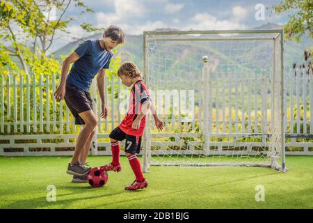 Kleiner niedlicher Junge in roter Fußballuniform und sein Trainer oder Vater spielt Fußball, Fußball auf dem Feld, im Freien. Aktives Kind macht Sport mit Stockfoto