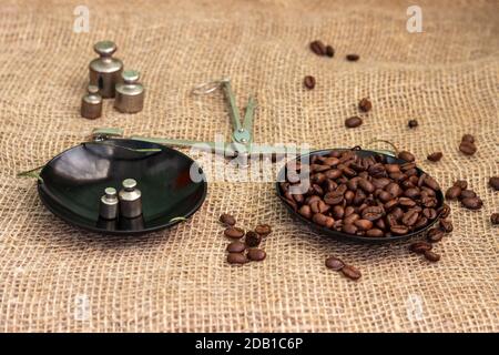Geröstete Kaffeebohnen, gewogen auf einer antiken Handwaage mit Gewichten, liegend auf einem mit Quappen bedeckten Tisch. Stockfoto