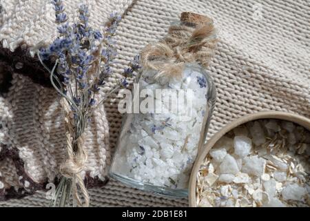 Eine Glasflasche Meersalz für ein entspannendes Bad Auf einem beigefarbenen Textil-Strickhintergrund neben einem kleinen Bouquet von Lavendel und einem Körperpeeling Stockfoto