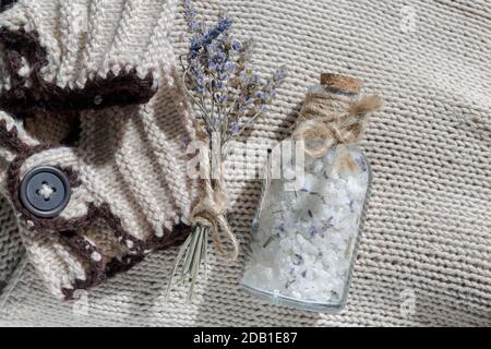 Eine Glasflasche Meersalz mit einem Korken für Ein entspannendes Bad auf beigefarbenem Textil-Strickhintergrund Zu einem kleinen Bouquet von Lavendel und einem Körperpeeling Stockfoto