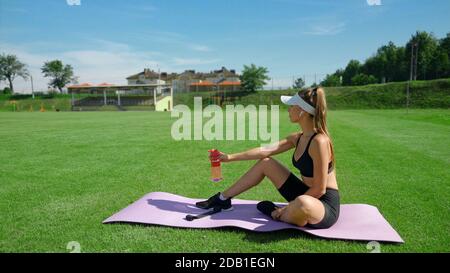 Seitenansicht der jungen fittigen Frau, die Flasche mit Wasser hält, auf der Matte sitzend, Stadionfeld im Sommer sonnigen Tag. Sportliche Mädchen tragen Sport-Outfit mit Ruhe auf grünem Gras. Konzept des Sports, Workout. Stockfoto