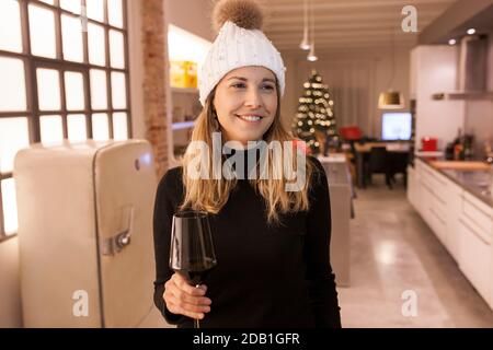 Schöne lächelnde Frau trägt weiße Beani und hält ein tasse Rotwein in der Küche mit weihnachtsbaum Hintergrund Stockfoto