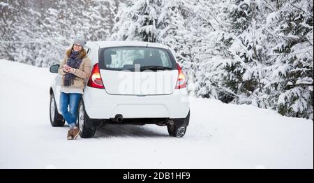 Frau mit Smartphone steht neben dem Auto auf einem Hintergrund Des schneebedeckten Winterwaldes Stockfoto