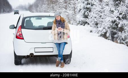 Frau mit Smartphone steht neben dem Auto auf einem Hintergrund Des schneebedeckten Winterwaldes Stockfoto