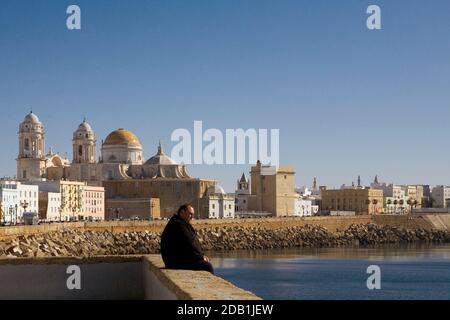 Mann sitzt an der Wand mit Blick auf den Ozean, mit dem Dom im Hintergrund: Calle Campo del Sur, Cádiz, Andalusien, Spanien Stockfoto