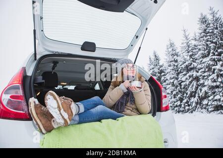 Frau sitzt im Kofferraum des Autos und hält eine Tasse heißen Tee in den Händen. Stockfoto