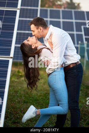Ein Mann küsst seine Geliebte vor dem Hintergrund einer Reihe von Solarzellen. Eine Frau mit langen Haaren genießt die Arme eines geliebten Menschen. Eine moderne Welt mit ständig weiterentwickelnden Technologien Stockfoto