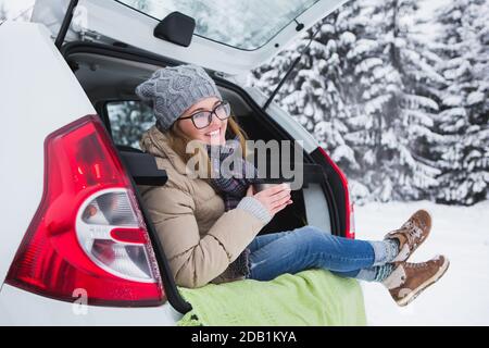 Frau sitzt im Kofferraum des Autos und hält eine Tasse heißen Tee in den Händen. Stockfoto