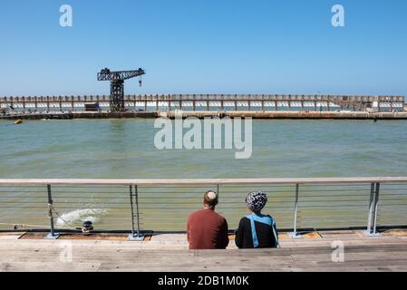 Jüdisches junges Paar (Mann mit Kippah und Frau mit Kopftuch), das sich am sonnigen Frühlingstag im Hafen von Tel Aviv, Israel, entspannt. Rückansicht; nicht erkennbar pe Stockfoto