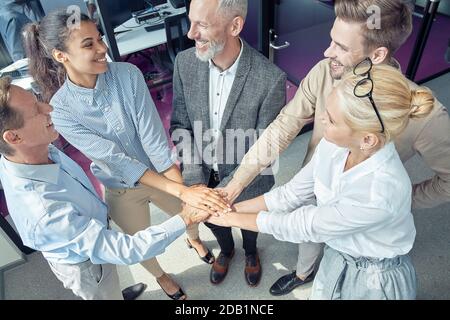 Gruppe von glücklichen motivierten Geschäftsleuten, die Hände zusammenlegen und lächeln, den Erfolg feiern, während sie im Büro stehen Stockfoto