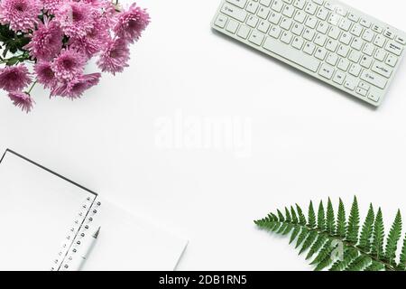 Notizbuch, Stift, Tastatur, Bouquet von rosa Chrysanthemen und Farnblätter auf weißem Hintergrund. Arbeitsplatz der Frau, Draufsicht, Kopierbereich. Stockfoto