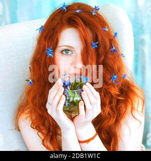 Ein nettes Rotschopf Mädchen sitzt in einem großen Stuhl mit einem Strauß von wilden blauen Schneeglöckchen in ihren Händen. Stockfoto