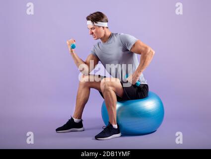 Entschlossener Millenial Sportler, der mit Hanteln trainiert, während er auf Fitness sitzt Ball auf flieder Studio Hintergrund Stockfoto