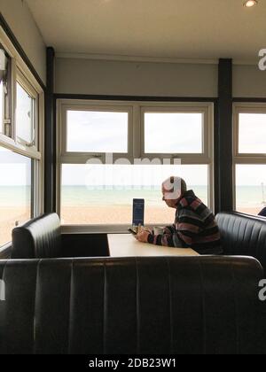 Ein Mann mittleren Alters sitzt allein in einem Café mit einer Tasse Kaffee SMS auf seinem Handy mit Blick auf das Meer in West Sussex, England, Großbritannien. Stockfoto
