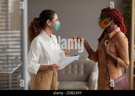 Taille oben Seitenansicht bei zwei fröhlichen Geschäftsfrauen tragen Masken Und mit Fäusten als kontaktlose Begrüßung im Büro Stockfoto