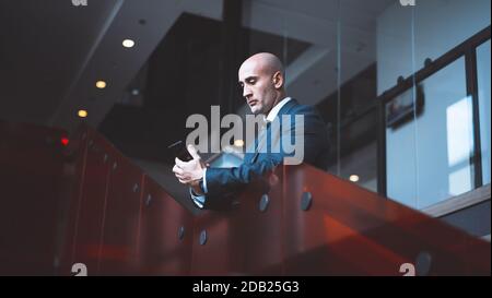 Serious kaukasischen Geschäftsmann hält Treffen online mit Telefon. Von unten betrachtet benutzt man ein Mobiltelefon, während man auf einer Treppe im Business Center steht Stockfoto