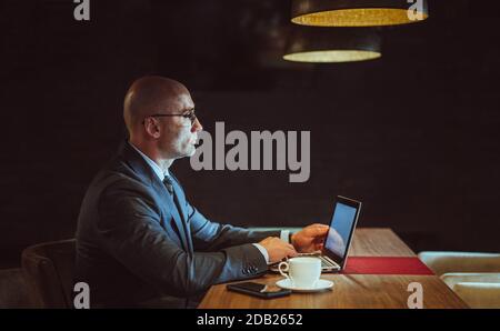 Im Business Center arbeitet ein Geschäftsmann am Computer. Seitenansicht des selbstbewussten kaukasischen Mann verwendet Laptop sitzen am Holztisch mit Kaffeetasse und mobile Stockfoto