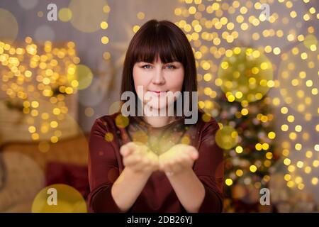 Weihnachten, Glück und Magie Konzept - Porträt der schönen Frau bläst Feenstaub von ihren Handflächen im dekorierten Wohnzimmer mit Weihnachtsbaum und Stockfoto