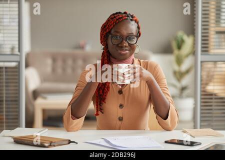 Vorderansicht Porträt einer jungen afroamerikanischen Frau, die Kaffeebecher hält und die Kamera anlächelt, während sie die Arbeit im Home Office genießt, Kopierraum Stockfoto