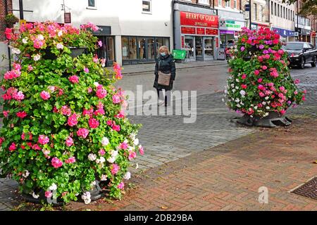 Frau trägt Covid 19 Coronavirus Gesichtsmaske allein entlang gehen In der Nähe verlassenen Brentwood Einkaufsstraße zwischen Blumenanzeige Essex England GB Stockfoto