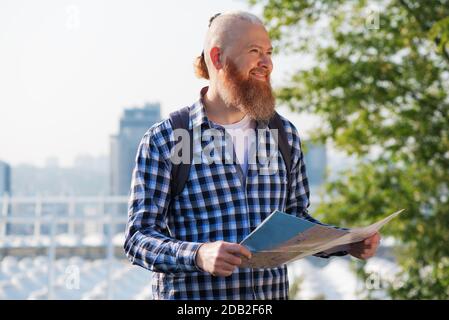 Lächelnder kaukasischer Hipster bärtiger Mann, der mit einem aktiven Urlaub zufrieden war. Lächelnder männlicher Reisender, der am Wochenende mit einer Karte durch die Stadt spazierenging Stockfoto