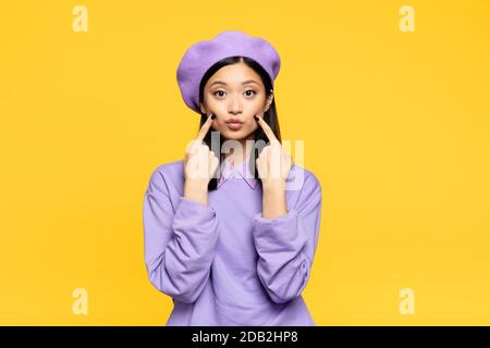 asiatische Frau in Baskenmütze berühren Wangen mit Fingern während pouting Lippen isoliert auf gelb Stockfoto