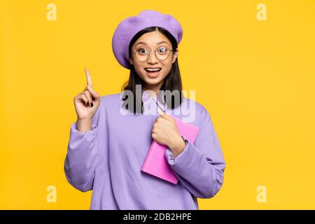 Aufgeregt asiatische Frau in Baskenmütze und Brillen halten Notebook und Zeigt mit Finger isoliert auf gelb Stockfoto