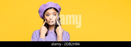 asiatische Frau in Baskenmütze berühren Wangen mit Fingern während Pouting Lippen isoliert auf gelb, Banner Stockfoto
