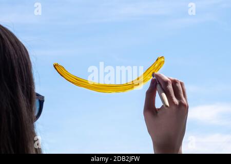 Weibliche Hand zeichnet ein lächelndes Gesicht mit Farbe auf dem Fenster gegen den blauen Himmel. Das Konzept von Glück, Gesundheit, Freude. Stockfoto