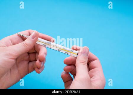 Nahaufnahme weibliche Hand hält ein Thermometer auf einem blauen Hintergrund. Thermometer zeigt eine hohe Körpertemperatur an. Stockfoto