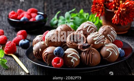 Schokoladentrüffelkugeln auf einem schwarzen Teller auf einem dunklen Holztisch mit frischen Beeren und Blumen im Hintergrund, horizontale Ansicht von oben Stockfoto