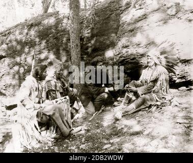 Drei Schwarzfuß-Indianer, die zeigen, wie man ein Feuer mit einem Bogen und einem Stock, Felsen und Bäume im Hintergrund zu starten. Stockfoto