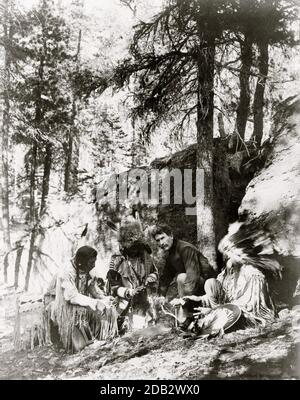 Ernest Thompson Seton, mit drei Schwarzfuß-Indianern, zeigt, wie man ein Feuer mit einem Bogen und einem Stock, Felsen und Bäumen im Hintergrund zu starten. Stockfoto