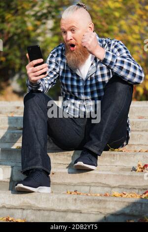 Aufgeregt überglücklich bärtigen Mann Blick auf Smartphone-Bildschirm sitzen auf Stufen im Freien Gefühl euphorisch Sieg feiern. Gewinner-Konzept Stockfoto