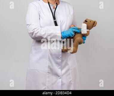 Weibliche Sanitäter halten braunen Teddybären mit Pfote in weißem Verband und hören Spielzeug mit Stethoskop, Konzept der Pädiatrie und Tierbehandlung Stockfoto