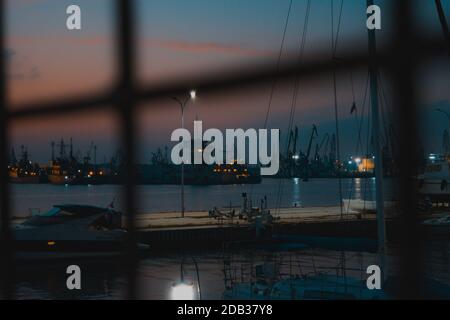 Atemberaubende Nachtansicht durch einen Metallzaun mit einer Yacht und einigen Schiffen im Hintergrund. Stockfoto