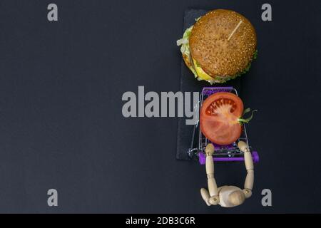 Figur eines kleinen Holzmanns, der einen Korb eines Supermarkts mit einer Tomate neben einem Hamburger schiebt. Top-Ansichten mit viel Platz Stockfoto